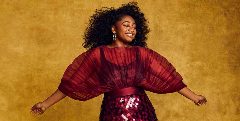 Colour photograph of singer Samara Joy smiling in a red dress against a yellow background.